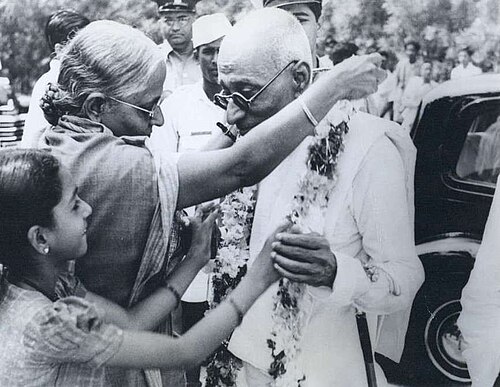 An elderly woman garlanding and blessing C. Rajagopalachari in a crowd, with a young girl beside her, as he stands wearing garlands next to a car.