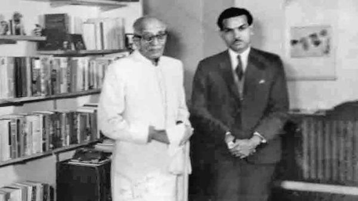 C. Rajagopalachari standing in a room lined with bookshelves, in white khadi, beside a younger man in a dark suit.